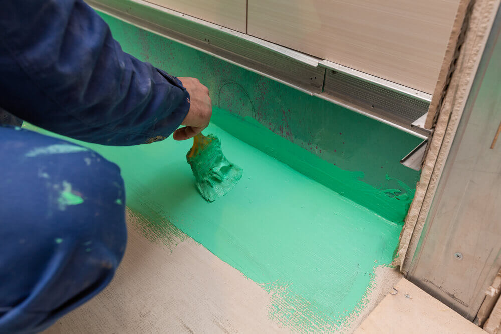 a tiler wearing blue overalls applying green waterproof paint to the edge of a bathroom floor