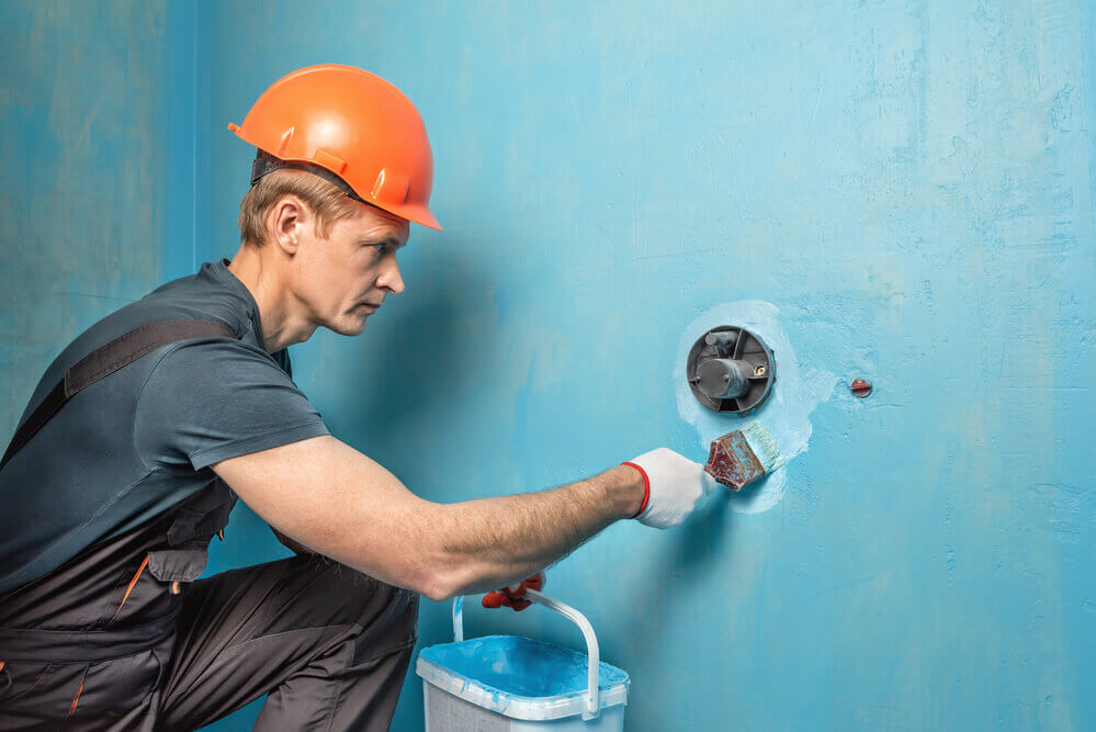 a tiler wearing a orange construction hard hat painting on blue waterproofing paint onto a pipe coming out of the wall